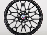 A black multi-spoke alloy wheel is displayed upright, featuring a BMW emblem in the center. The intricate design sits against a plain, light gray backdrop.