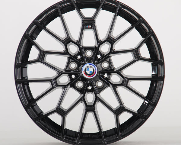 A black multi-spoke alloy wheel is displayed upright, featuring a BMW emblem in the center. The intricate design sits against a plain, light gray backdrop.