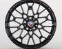 A black multi-spoke alloy wheel is displayed upright, featuring a BMW emblem in the center. The intricate design sits against a plain, light gray backdrop.