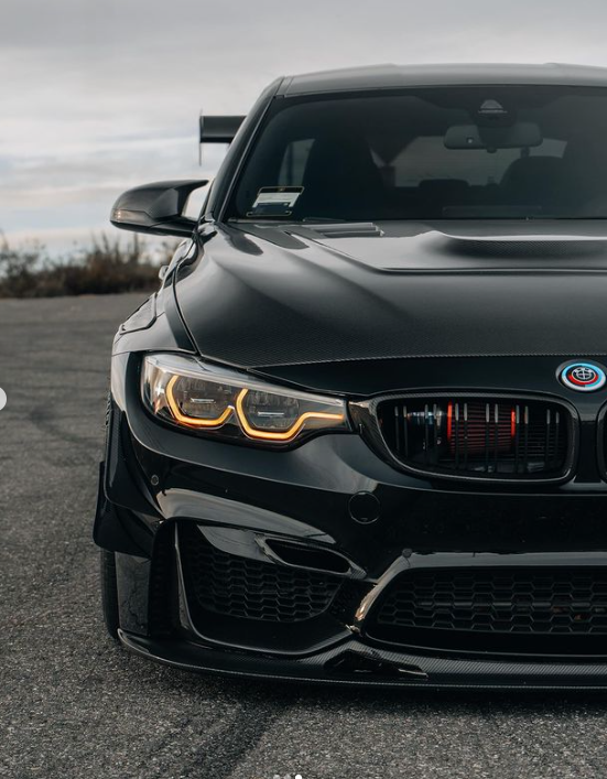 A sleek black car with glowing headlights is parked on a paved surface, with a cloudy sky in the background. The car's design is sporty and aggressive.