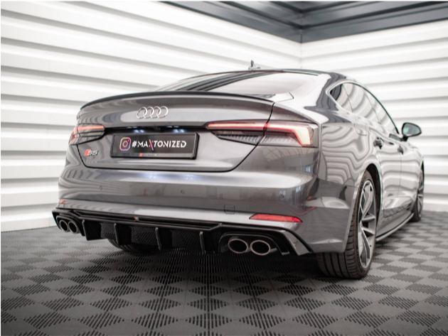 A sleek gray car, positioned in a showroom with white walls and patterned floor, has a visible rear badge marked "S5." The license plate reads "#MAXTONIZED."