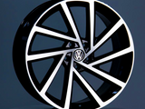 A black and silver car wheel with a geometric spoke design, displaying a Volkswagen logo in the center, is positioned upright on a reflective surface within a gray environment.