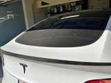 A white Tesla car is parked in a garage. The view shows the rear window and trunk, highlighted by a carbon fiber spoiler. Shelving with various items is visible in the background.