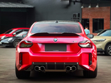 A red BMW M2 is parked on a paved surface, featuring quad exhausts, within an urban setting with several other cars and a building visible in the background.