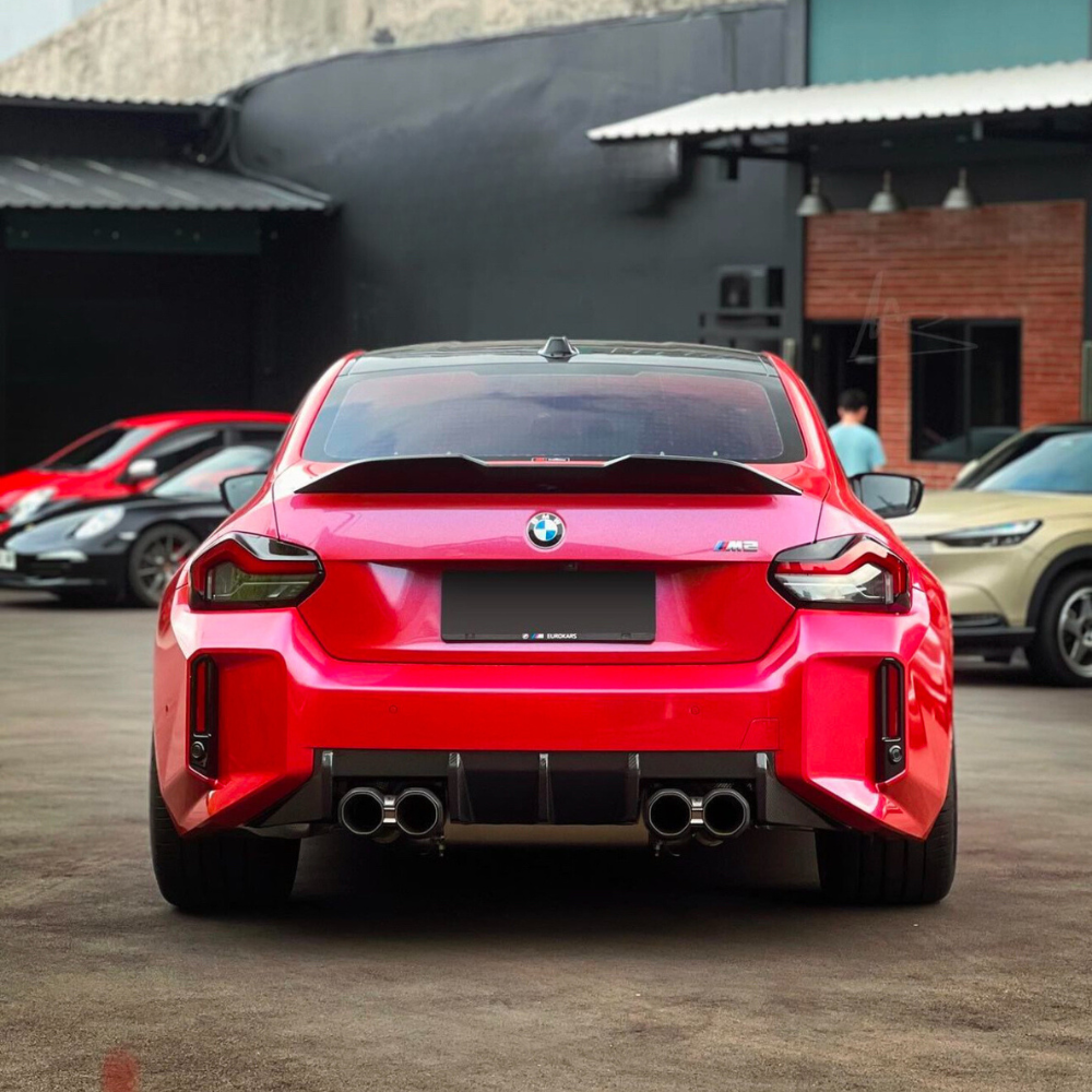 A red BMW M2 is parked on a paved surface, featuring quad exhausts, within an urban setting with several other cars and a building visible in the background.
