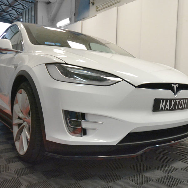 A white car with a Tesla logo is parked indoors on a checkered floor, displaying the license plate "MAXTON." The room features a high ceiling and bright lighting.