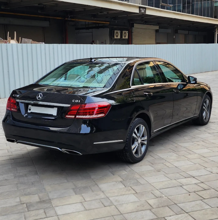 A black Mercedes-Benz E250 CDI is parked on a tiled surface, adjacent to a blue metal fence, with industrial buildings in the background.