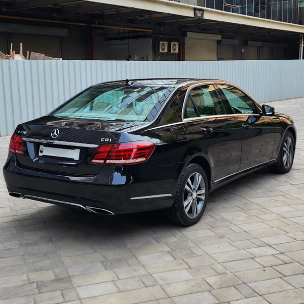 A black Mercedes-Benz E250 CDI is parked on a tiled surface, adjacent to a blue metal fence, with industrial buildings in the background.