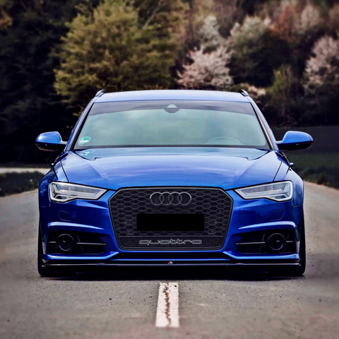 A blue Audi car is parked on a rural road, facing forward. "quattro" is written on the grille. Trees and foliage line the background, creating a natural setting.