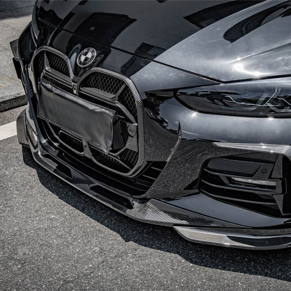 A front view of a sleek black car with a prominent grille. The vehicle is parked on a paved street, reflecting light and nearby structures.