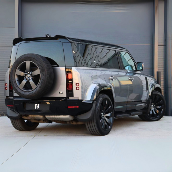 A silver SUV with a rear-mounted spare tire is parked on a light-colored pavement in front of a grey industrial building. The vehicle features dark-tinted windows and large black wheels.
