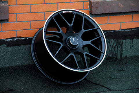 A black alloy wheel with a silver edge rests against an orange brick wall on a dark surface. The central cap features a three-pointed star logo and "AMG" is printed above.