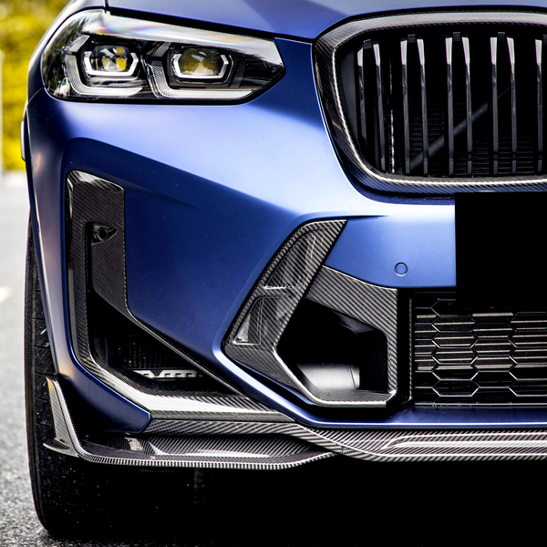 The close-up of a blue car front highlights a sleek headlight and sporty grille with carbon fiber detailing, parked on a road with a blurred background.