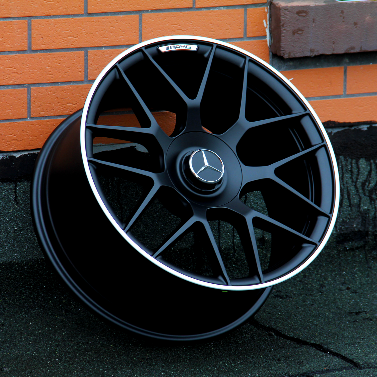 A black alloy wheel with the Mercedes-Benz logo and "AMG" text rests against a textured orange brick wall on a gray surface.