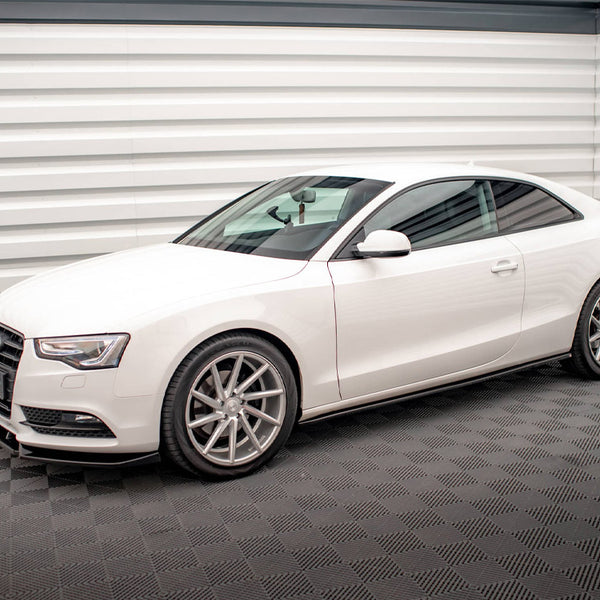 A white Audi coupe is parked, showcasing its sleek design with silver alloy wheels, in a well-lit room with textured gray flooring and horizontal white paneling on the walls.