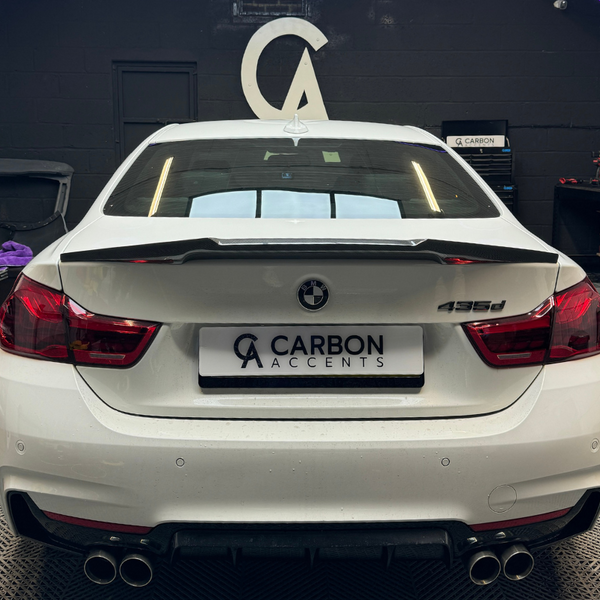 A white BMW 435d with a custom spoiler is parked on a vehicle lift inside an automotive workshop. The license plate reads "CARBON ACCENTS" with a logo above.