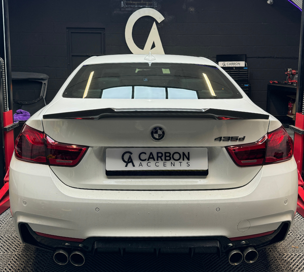 A white BMW 435d with a custom spoiler is parked on a vehicle lift inside an automotive workshop. The license plate reads "CARBON ACCENTS" with a logo above.