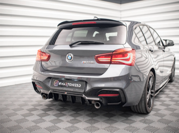 A gray BMW hatchback is parked on a patterned floor in a well-lit, corner room with white walls. The car features dual exhausts and red taillights. Text: "#MAXTONIZED".