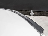 A carbon fiber car spoiler on a white vehicle is shown close-up, situated in an outdoor setting near a concrete wall and metal barrier post.