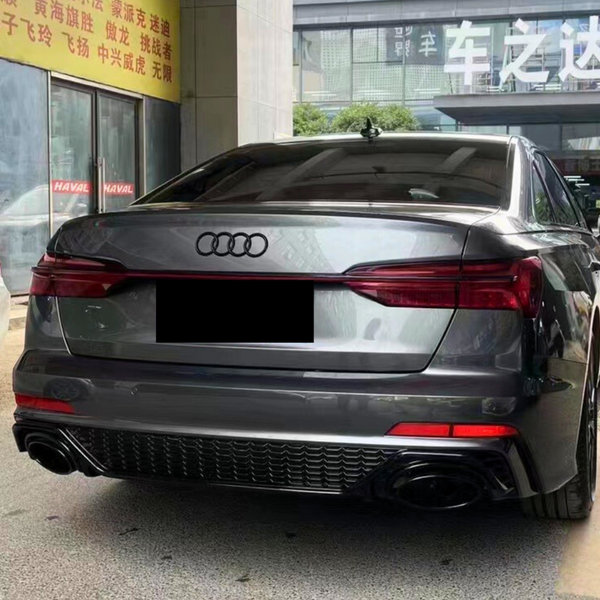 A gray Audi car is parked near a building with signs. Chinese text on a yellow sign and white text on glass are visible, and the ground is paved.