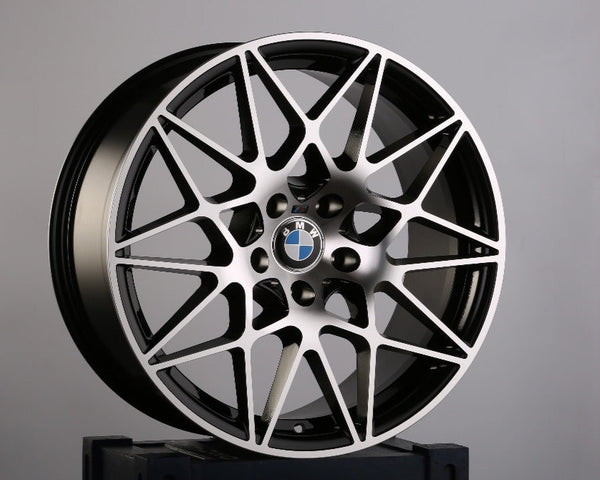 A silver and black alloy wheel with intricate geometric spokes rests on a blue crate with partial text "GA" and a visible UK flag. BMW logo is at the center.