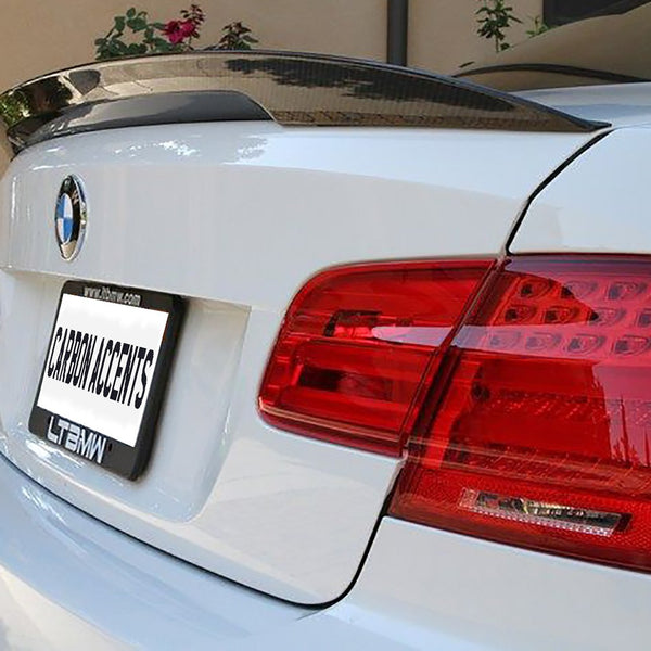 A white car features a carbon fiber spoiler and red tail lights. The rear license plate shows the text "CARBON ACCENTS." It's parked in a driveway with plants nearby.