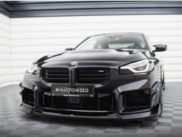 A black sports car with a sleek, aerodynamic design is parked on a textured floor in a well-lit garage. The license plate reads "#MAXTONIZED Dealer BMW M//Cars."