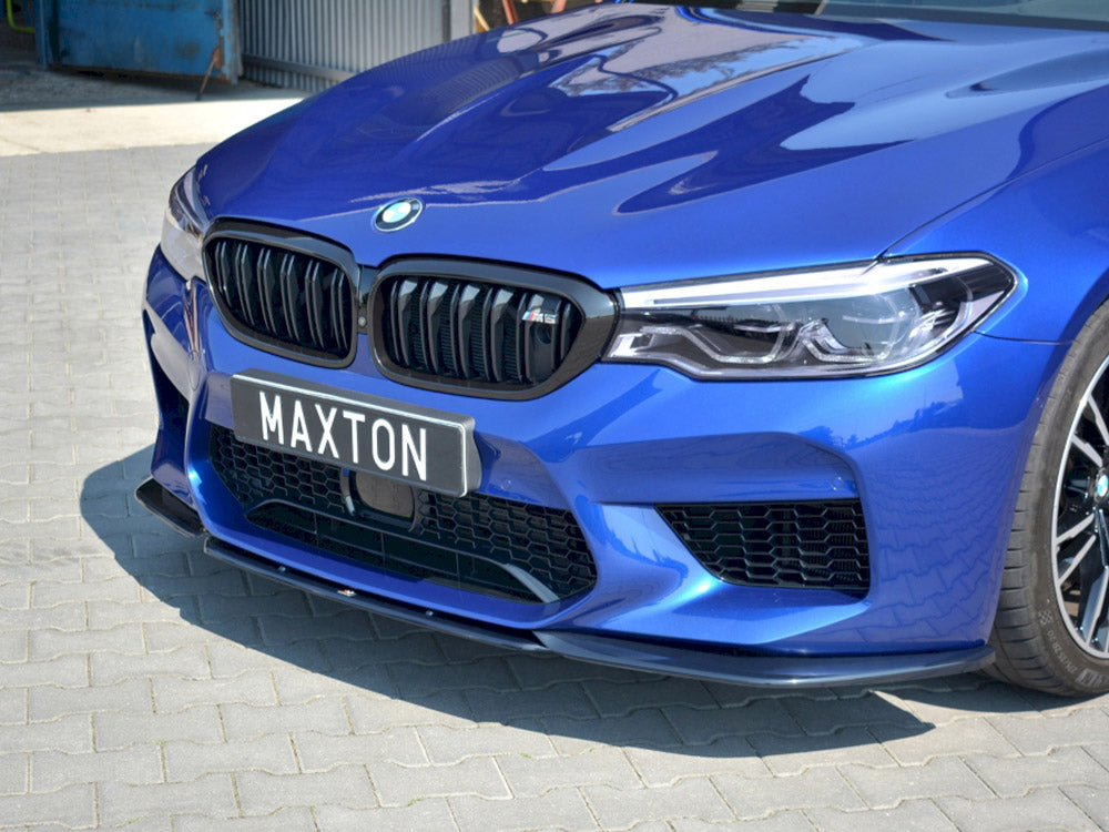 A blue car with a prominent grille and headlights, parked on a paved surface. The license plate reads "MAXTON." It is parked near a corrugated metal structure and industrial environment.