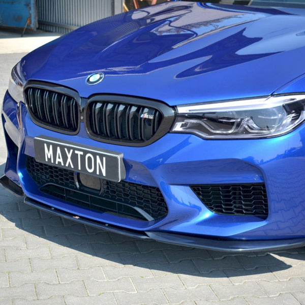 A blue car with a prominent grille and headlights, parked on a paved surface. The license plate reads "MAXTON." It is parked near a corrugated metal structure and industrial environment.