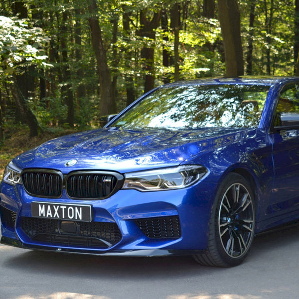 A blue car with sleek design stands parked on a shaded road, surrounded by dense green forest. The license plate reads "MAXTON."