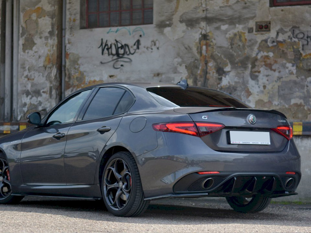 A sleek gray car is parked on a paved surface, its rear facing a weathered, graffiti-marked wall with peeling paint.