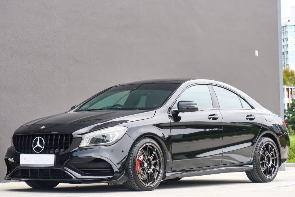 A sleek black car is parked on a smooth surface, featuring shiny black wheels. It is positioned against a plain gray wall, with buildings visible in the background.