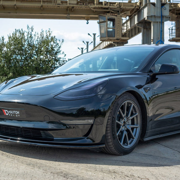 A sleek black car is parked on a concrete surface near industrial structures and greenery. The front bumper displays a "Maxton Design" logo.