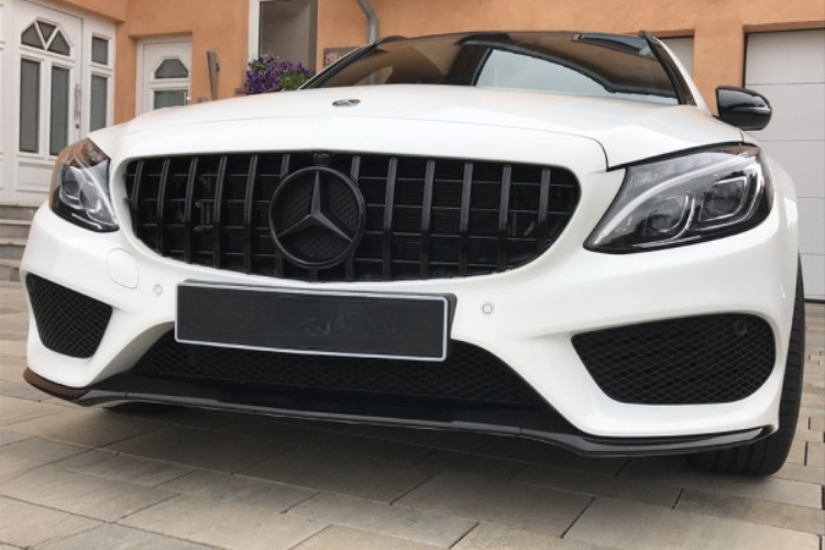 A white Mercedes-Benz car parked on paved ground, displaying a prominent grille with a logo. The background includes a peach-colored building with windows and a door.