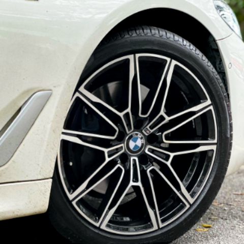 A car's front wheel is stationary, featuring a multi-spoke design with a BMW emblem, against a paved road background. The car is white with visible tire treads.
