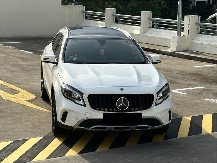 A white Mercedes-Benz is parked on a rooftop parking lot, positioned diagonally on yellow and black striped markings, surrounded by concrete barriers and railings under a clear sky.