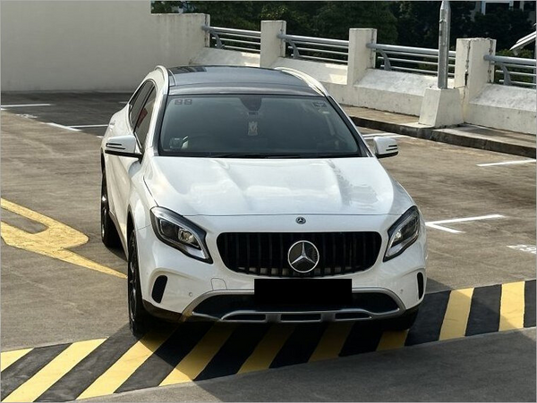 A white Mercedes-Benz is parked on a rooftop parking lot, positioned diagonally on yellow and black striped markings, surrounded by concrete barriers and railings under a clear sky.