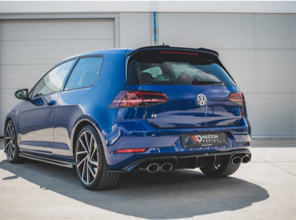 A blue Volkswagen car is parked in an industrial area with closed garage doors. It features dual exhausts and alloy wheels. The license plate reads "MAXTON DESIGN."