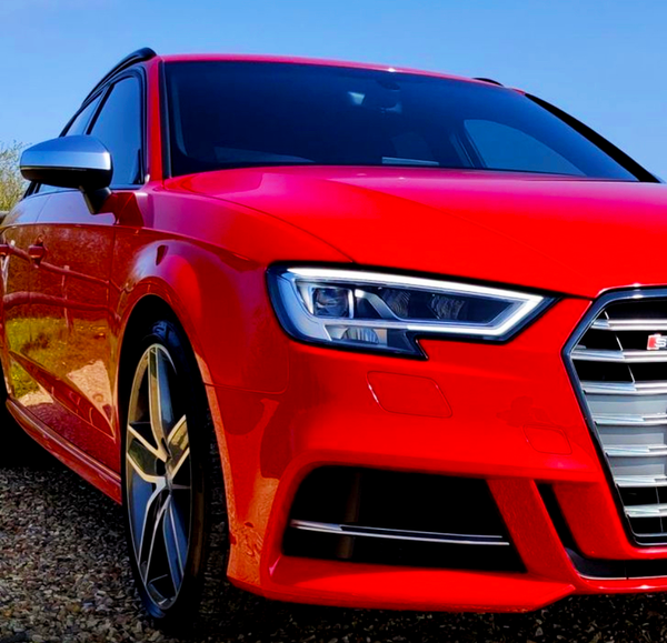 A red sports car with sleek headlights is parked on gravel, under a clear blue sky. The car's distinctive grille and angular design suggest high performance and modern styling.