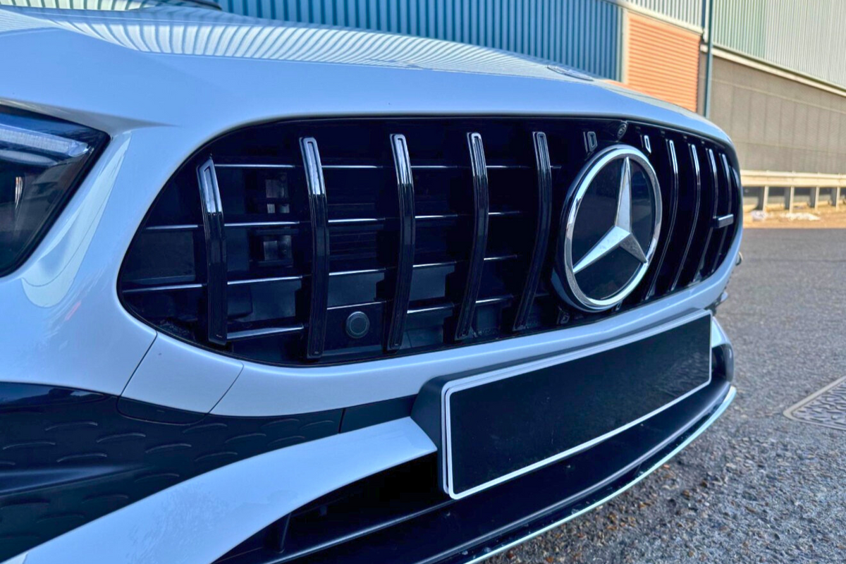 A white car features a prominent black grille with a silver emblem, parked on asphalt near blue and orange industrial buildings.