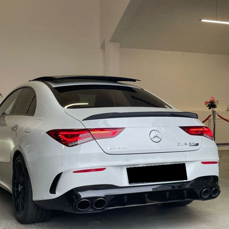 A white Mercedes-Benz CLA 45 AMG is parked indoors, showcasing distinct rear features, including dual exhausts and AMG badging. The setting includes a minimal, well-lit garage with decorative flowers.