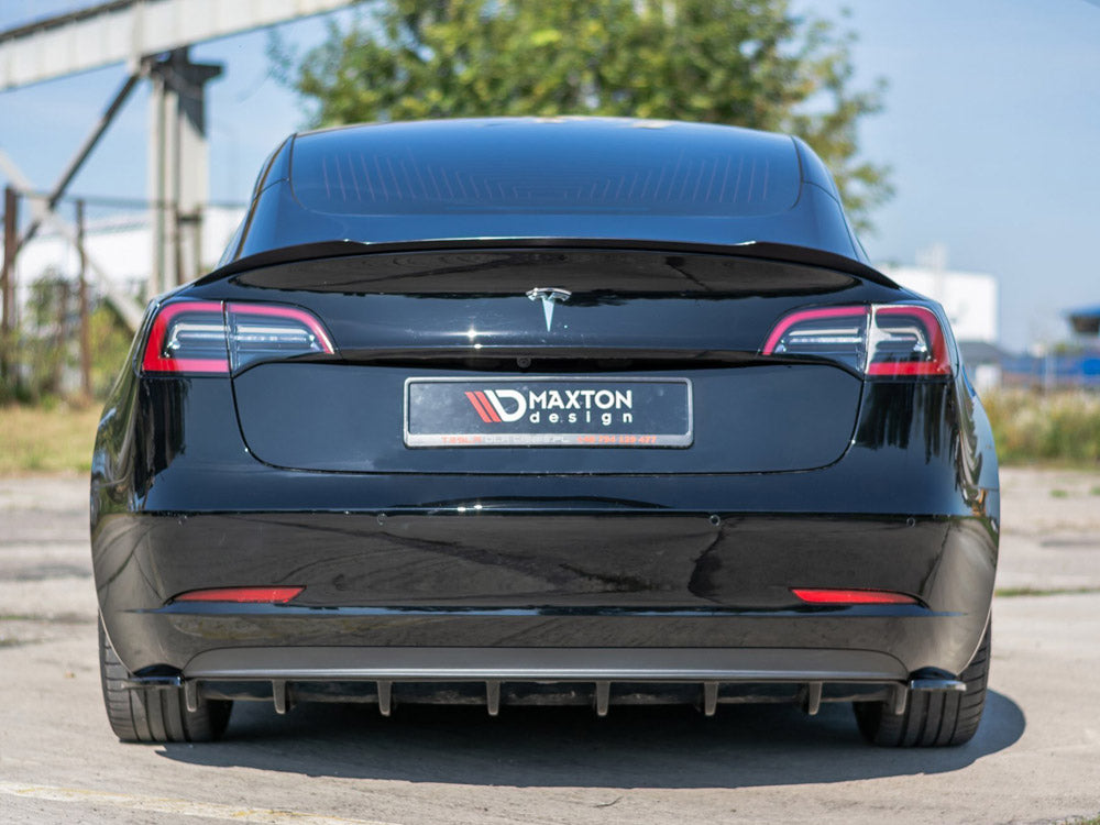 A black car, viewed from the rear, displays a license plate with "MAXTON design" text. It is parked on a concrete surface next to a fence and greenery under a clear sky.