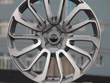 A silver alloy car wheel with intricate spoke design stands upright on a concrete block. The central hub features the text 