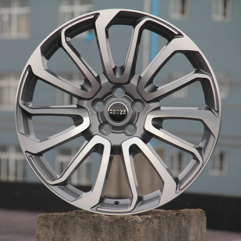 A silver alloy car wheel with intricate spoke design stands upright on a concrete block. The central hub features the text "RANGE ROVER." In the background, blurred urban buildings are visible.