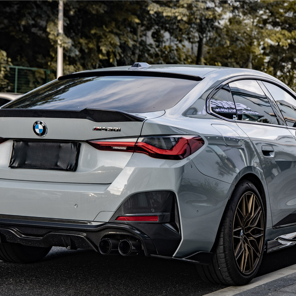 A silver BMW M440i parked on a street, featuring a sleek rear design and sports exhaust. Surrounding area includes other cars and trees lining the road.