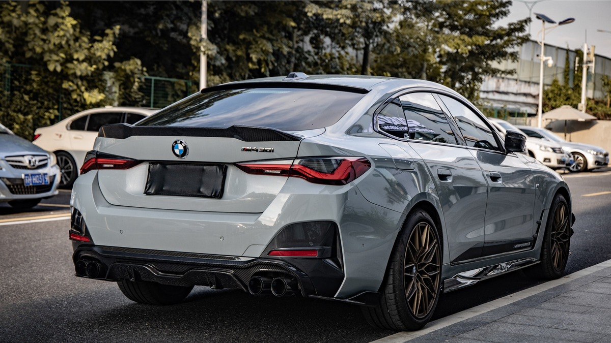 A silver BMW M440i parked on a street, featuring a sleek rear design and sports exhaust. Surrounding area includes other cars and trees lining the road.