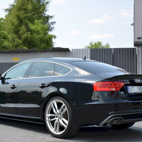 A black Audi car is parked outdoors on a paved surface with trees and a corrugated metal fence in the background. License plate details are blurred.