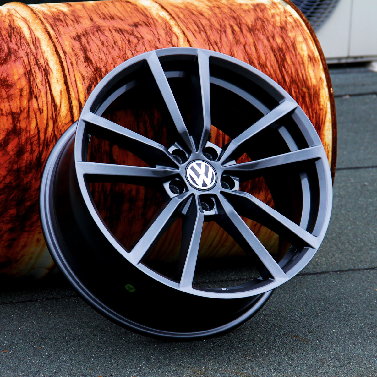 A black Volkswagen alloy wheel rests against a large orange cylinder with a wood-like texture on a gray surface.