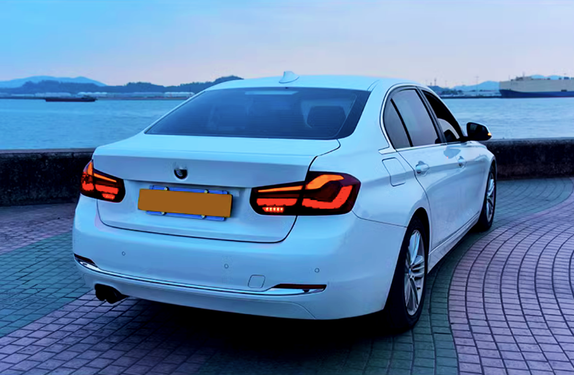 A white sedan with illuminated taillights is parked on a patterned pavement beside a waterfront, with distant hills and a ship visible across a calm body of water.