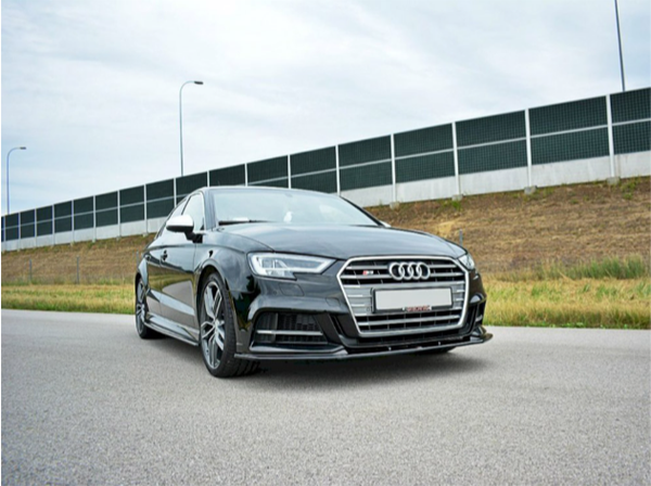 A black Audi car is parked on a road, with a grassy embankment and noise-barrier fencing in the background under a cloudy sky.